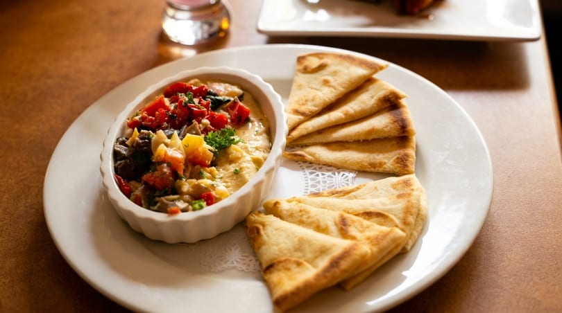 Hummus topped with diced tomatoes and vegetables, served with pita bread on a white plate.