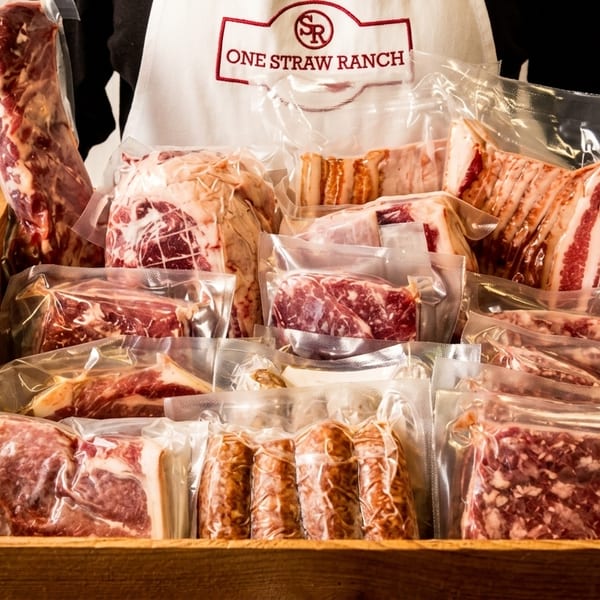Hands holding a wooden crate filled with various vacuum-sealed meat products from One Straw Ranch.