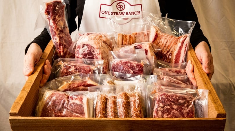 Hands holding a wooden crate filled with various vacuum-sealed meat products from One Straw Ranch.