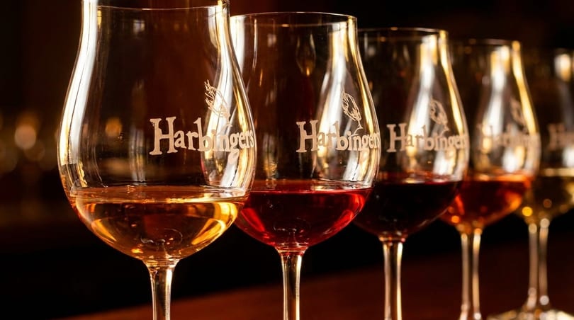 A row of wine glasses filled with red, rosé, and white wine, branded "Harbingers".
