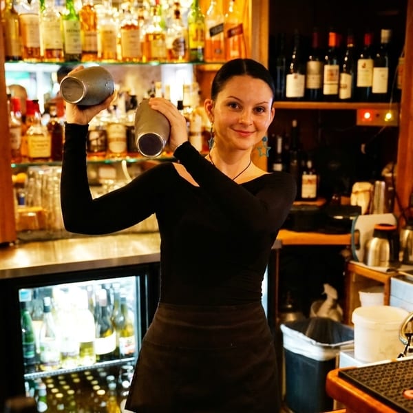 Smiling bartender in black shirt and apron shaking a cocktail at a well-stocked bar.