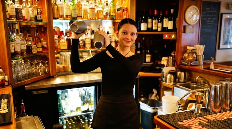 Smiling bartender in black shirt and apron shaking a cocktail at a well-stocked bar.