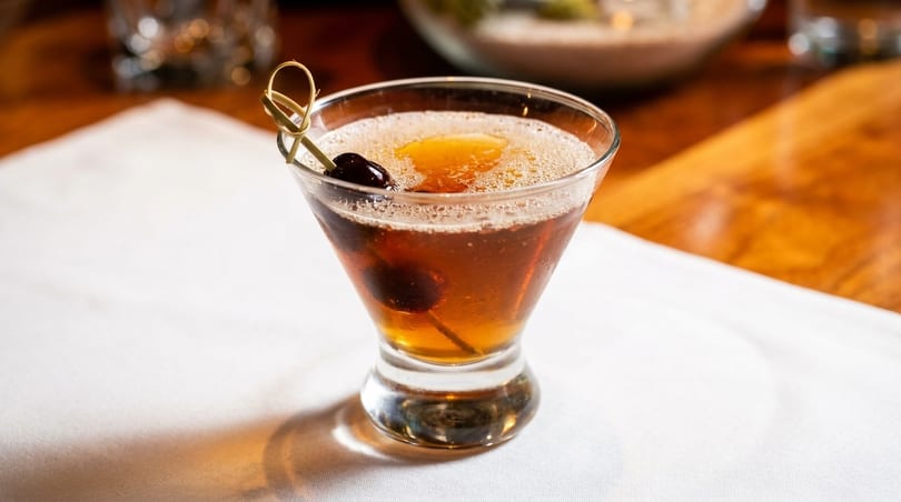 A Manhattan cocktail in a martini glass, garnished with a cherry on a pick, on a white tablecloth.