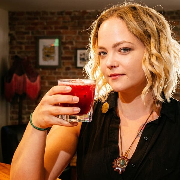 Blonde woman holding a red cocktail, with a stained-glass window and brick wall in the background.