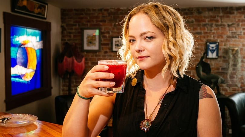 Blonde woman holding a red cocktail, with a stained-glass window and brick wall in the background.