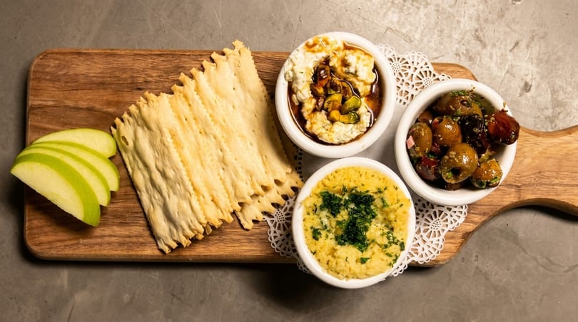 A wooden board with crackers, apple slices, and three small bowls of dips and olives.