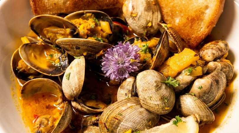 Steamed clams in a rich broth with herbs, lemon, and toasted bread, garnished with a purple flower.