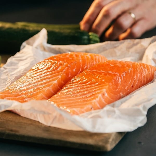 Two fresh salmon fillets on parchment paper, with carrots and zucchini in the background.