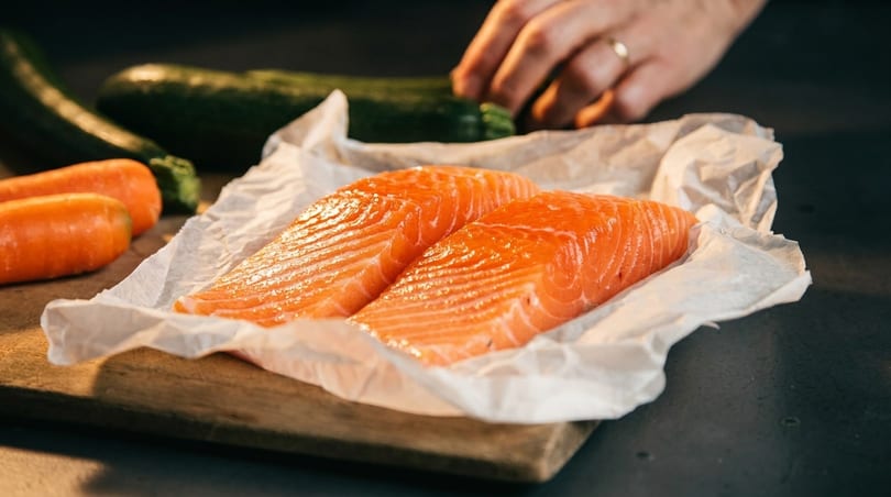 Two fresh salmon fillets on parchment paper, with carrots and zucchini in the background.