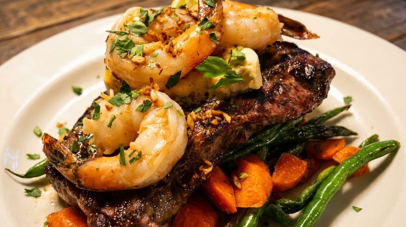 Grilled steak and shrimp with green beans and carrots on a white plate.