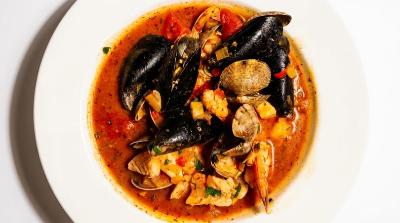 A white bowl filled with seafood cioppino, featuring mussels, clams, shrimp, and fish in a rich tomato broth.