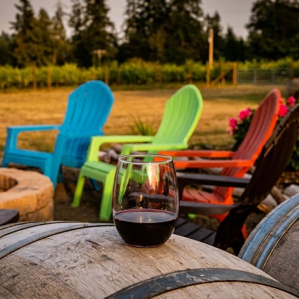 A glass of red wine on a barrel, with colorful Adirondack chairs and a fire pit in a vineyard.